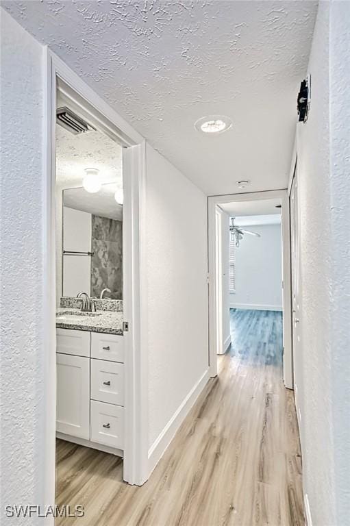 hallway featuring visible vents, baseboards, a textured ceiling, a sink, and light wood-style flooring