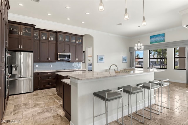 kitchen featuring tasteful backsplash, hanging light fixtures, a center island with sink, ornamental molding, and stainless steel appliances