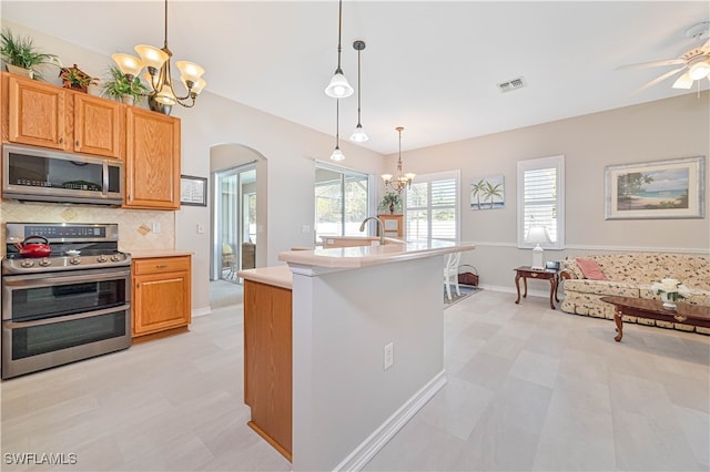 kitchen with appliances with stainless steel finishes, ceiling fan with notable chandelier, backsplash, decorative light fixtures, and a center island with sink