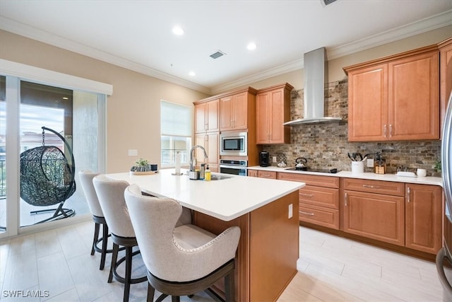 kitchen with a sink, light countertops, wall chimney range hood, appliances with stainless steel finishes, and a center island with sink