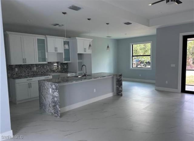 kitchen with stone countertops, a center island with sink, white cabinets, pendant lighting, and backsplash