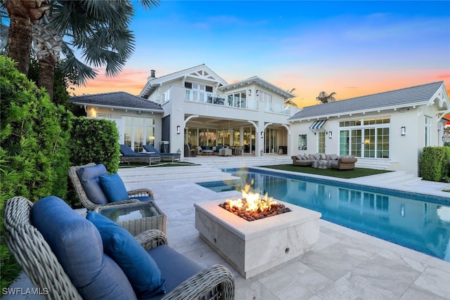 back house at dusk featuring a balcony, a patio, and an outdoor living space with a fire pit