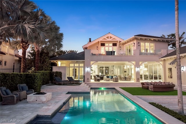 back house at dusk with a fenced in pool, an outdoor living space with a fire pit, a patio, and a balcony