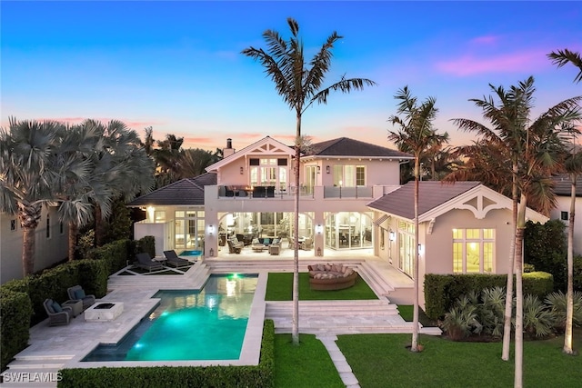 back house at dusk with a balcony, a yard, a pool with hot tub, a patio, and an outdoor living space with a fire pit