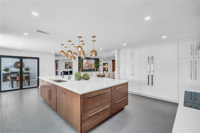 kitchen with an island with sink, sink, light tile patterned flooring, decorative light fixtures, and white cabinetry