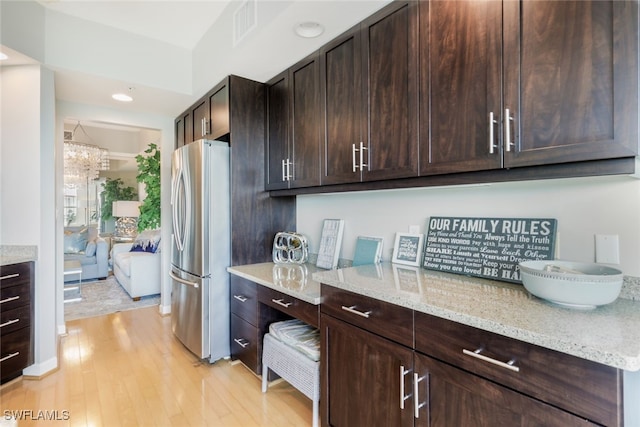 kitchen with stainless steel fridge, dark brown cabinets, light stone countertops, a chandelier, and light hardwood / wood-style flooring