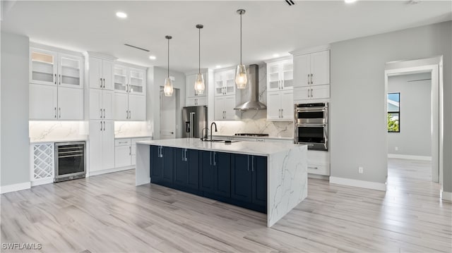 kitchen with beverage cooler, appliances with stainless steel finishes, an island with sink, wall chimney exhaust hood, and white cabinets