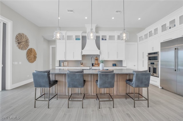 kitchen featuring hanging light fixtures, a center island with sink, light stone counters, premium range hood, and stainless steel appliances