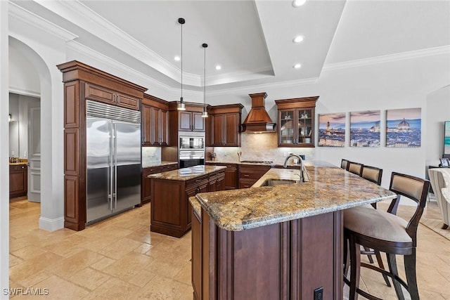 kitchen featuring custom range hood, glass insert cabinets, decorative light fixtures, built in appliances, and a sink