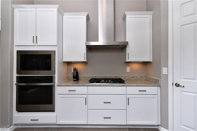 kitchen featuring tile patterned floors, wall chimney exhaust hood, light stone countertops, appliances with stainless steel finishes, and white cabinetry