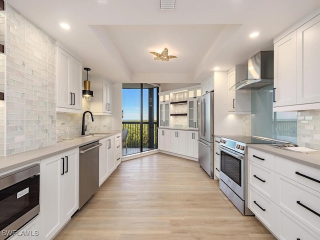 kitchen with appliances with stainless steel finishes, light wood-type flooring, sink, wall chimney range hood, and white cabinets