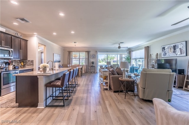 kitchen with dark brown cabinets, stainless steel appliances, a center island with sink, crown molding, and light wood-type flooring