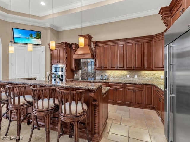 kitchen with pendant lighting, custom exhaust hood, a center island with sink, built in appliances, and stone countertops