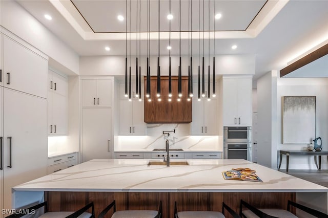 kitchen with light stone countertops, a tray ceiling, a large island with sink, decorative light fixtures, and white cabinetry