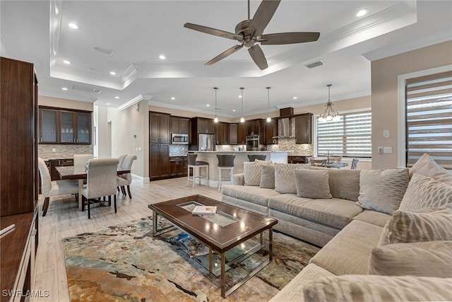 living room featuring crown molding, a tray ceiling, light wood-type flooring, and ceiling fan with notable chandelier