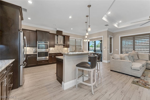 kitchen with a kitchen island, wall chimney exhaust hood, stainless steel appliances, a kitchen bar, and light stone counters