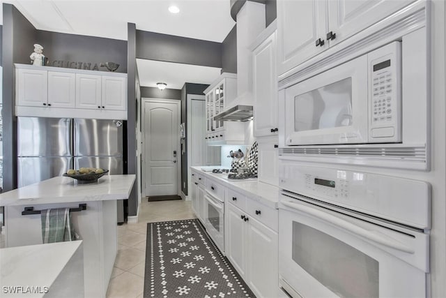 kitchen with wall chimney range hood, white cabinetry, light tile patterned floors, and white appliances