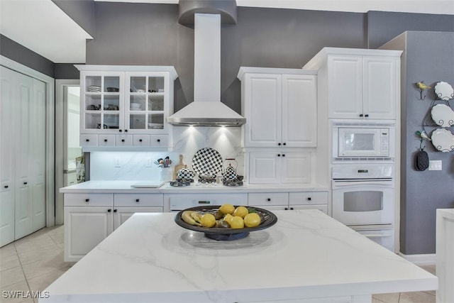 kitchen with wall chimney range hood, white appliances, white cabinetry, backsplash, and light stone counters