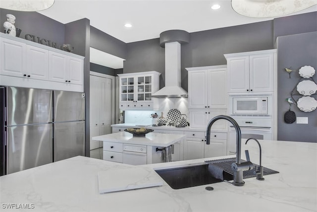 kitchen with stainless steel refrigerator, wall chimney exhaust hood, light stone counters, and white cabinets