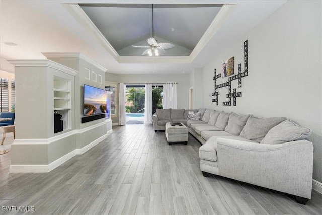 living room featuring ornamental molding, built in shelves, a raised ceiling, ceiling fan, and light hardwood / wood-style flooring