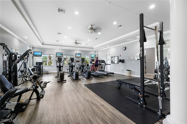 exercise room featuring ornate columns, wood-type flooring, ceiling fan, crown molding, and a raised ceiling