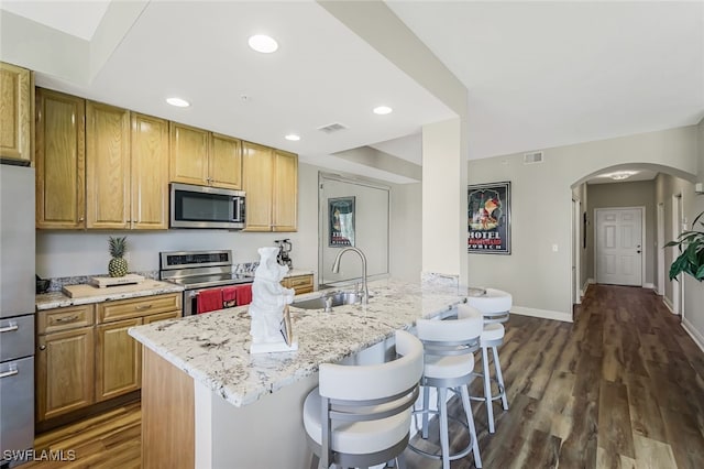 kitchen with an island with sink, appliances with stainless steel finishes, a kitchen breakfast bar, dark hardwood / wood-style floors, and light stone counters