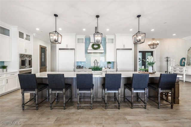 kitchen featuring stainless steel oven, a chandelier, custom exhaust hood, a large island with sink, and light countertops