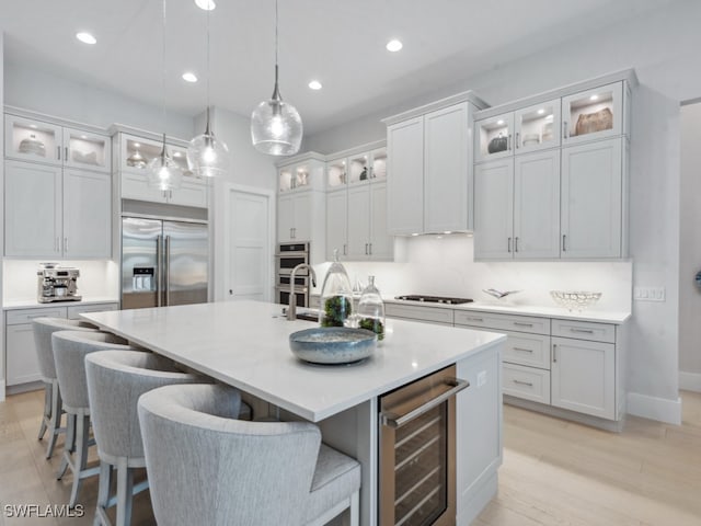 kitchen featuring wine cooler, appliances with stainless steel finishes, a kitchen island with sink, light wood-type flooring, and pendant lighting