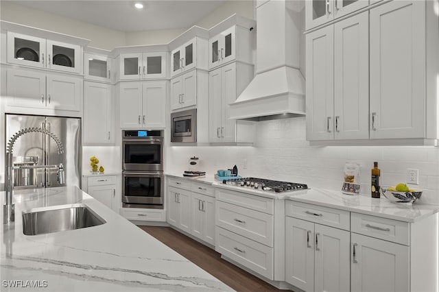 kitchen featuring decorative backsplash, appliances with stainless steel finishes, white cabinets, a sink, and premium range hood