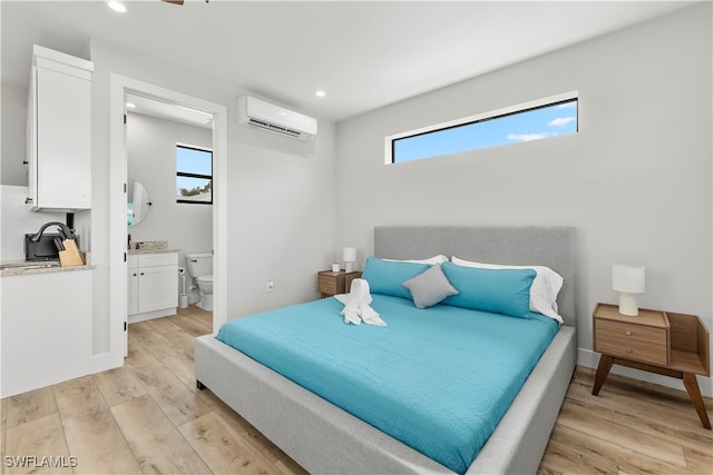 bedroom featuring a wall unit AC, multiple windows, ensuite bath, and light wood-type flooring