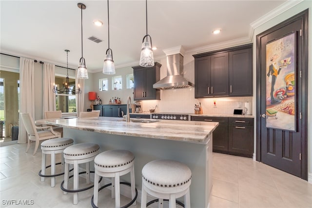 kitchen featuring ornamental molding, wall chimney exhaust hood, a kitchen island with sink, sink, and decorative light fixtures