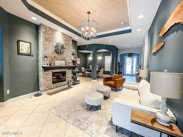 living room with an inviting chandelier, a tray ceiling, ornamental molding, a stone fireplace, and tile patterned floors