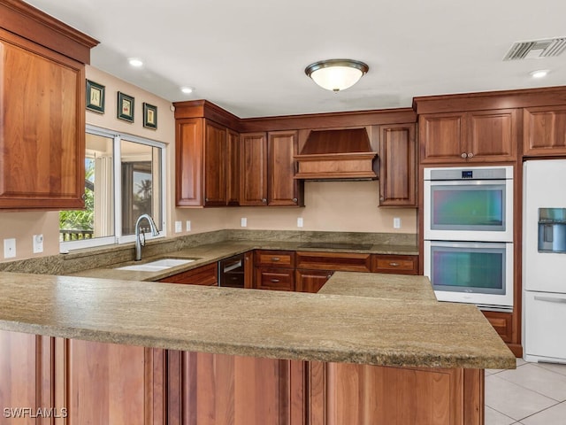 kitchen featuring kitchen peninsula, double wall oven, custom range hood, sink, and white refrigerator with ice dispenser