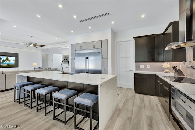 kitchen with a kitchen island with sink, a kitchen breakfast bar, wall chimney range hood, sink, and appliances with stainless steel finishes