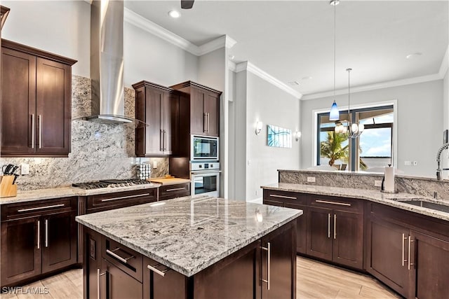 kitchen with wall chimney range hood, sink, hanging light fixtures, stainless steel appliances, and a kitchen island