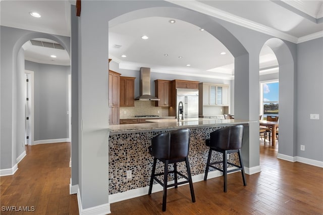 kitchen featuring ornamental molding, stainless steel built in fridge, dark wood-type flooring, wall chimney range hood, and decorative backsplash