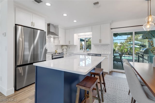 kitchen with hanging light fixtures, light hardwood / wood-style floors, stainless steel appliances, wall chimney exhaust hood, and white cabinets