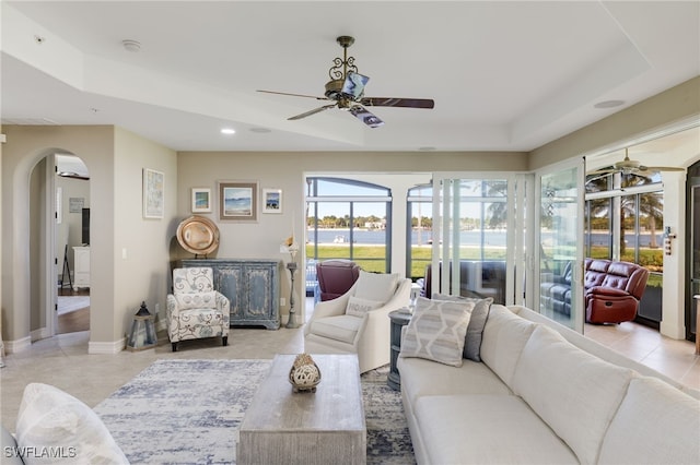 living room featuring light tile patterned floors, arched walkways, baseboards, ceiling fan, and a tray ceiling