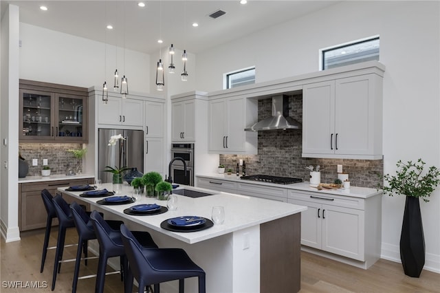 kitchen with tasteful backsplash, wall chimney range hood, stainless steel appliances, light stone counters, and a kitchen island with sink