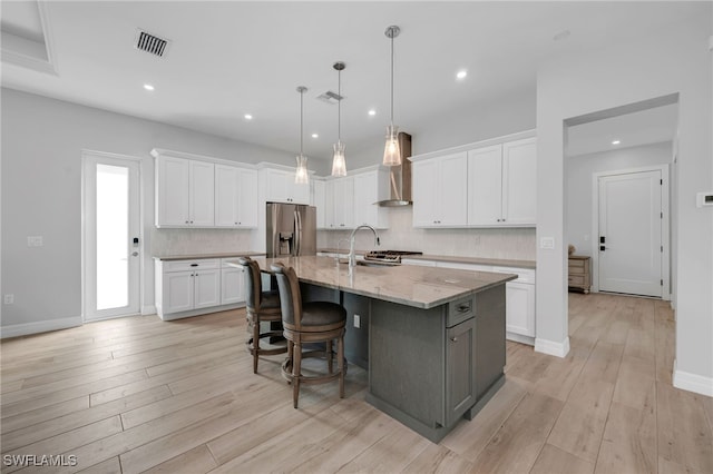 kitchen with white cabinetry, light stone countertops, wall chimney range hood, stainless steel fridge with ice dispenser, and an island with sink