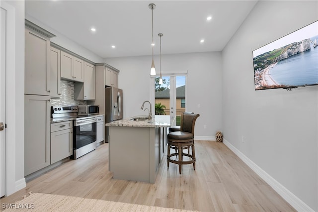 kitchen featuring light stone countertops, appliances with stainless steel finishes, light wood-type flooring, gray cabinetry, and a kitchen island with sink