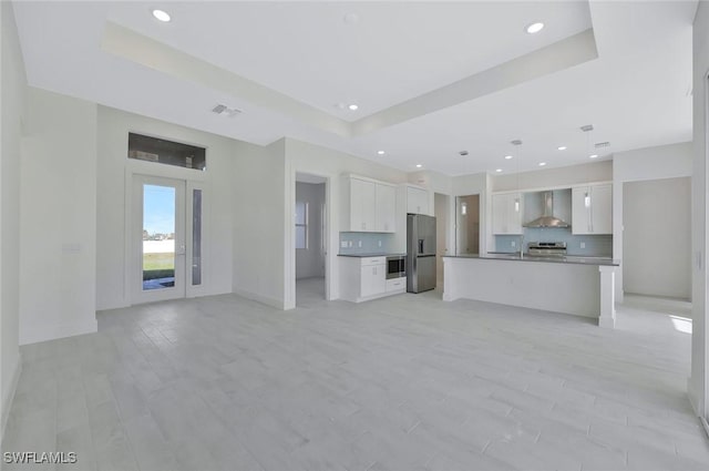 kitchen with a raised ceiling, appliances with stainless steel finishes, open floor plan, white cabinetry, and wall chimney exhaust hood