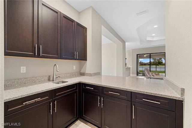 kitchen featuring sink, light stone counters, dark brown cabinetry, a tray ceiling, and kitchen peninsula