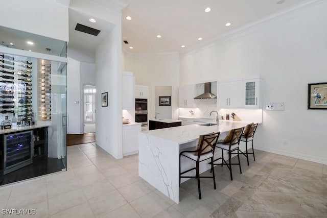 kitchen with wall chimney exhaust hood, a towering ceiling, double oven, a sink, and a kitchen breakfast bar