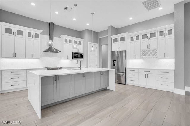 kitchen featuring stainless steel appliances, wall chimney exhaust hood, pendant lighting, white cabinets, and a kitchen island with sink