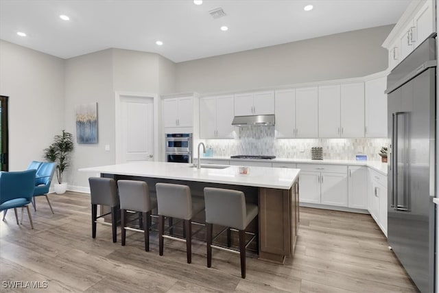 kitchen featuring decorative backsplash, appliances with stainless steel finishes, white cabinetry, an island with sink, and under cabinet range hood