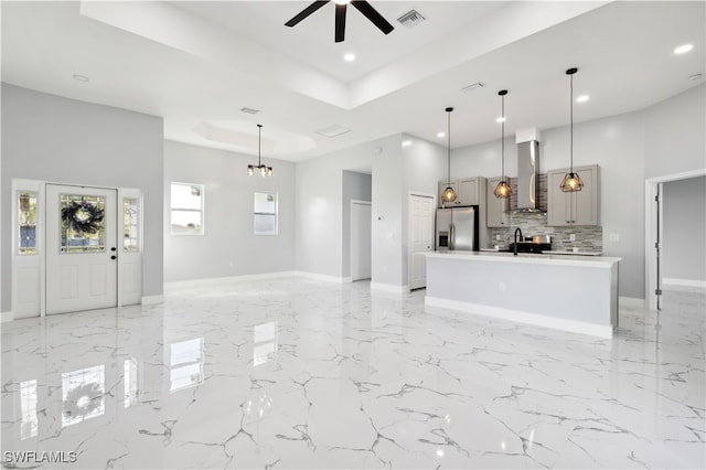 kitchen featuring a kitchen island with sink, gray cabinets, stainless steel fridge, pendant lighting, and wall chimney range hood