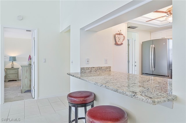 kitchen featuring kitchen peninsula, stainless steel fridge, white cabinetry, light stone countertops, and a kitchen bar