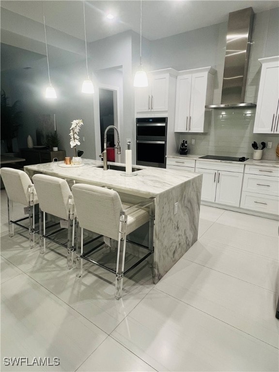 kitchen featuring wall chimney range hood, white cabinets, hanging light fixtures, light stone countertops, and a kitchen island with sink