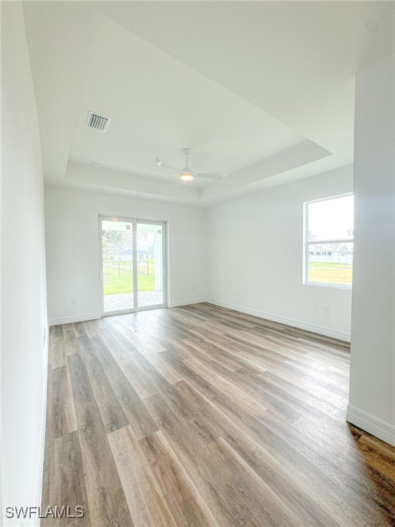 empty room with a wealth of natural light, ceiling fan, light wood-type flooring, and a raised ceiling